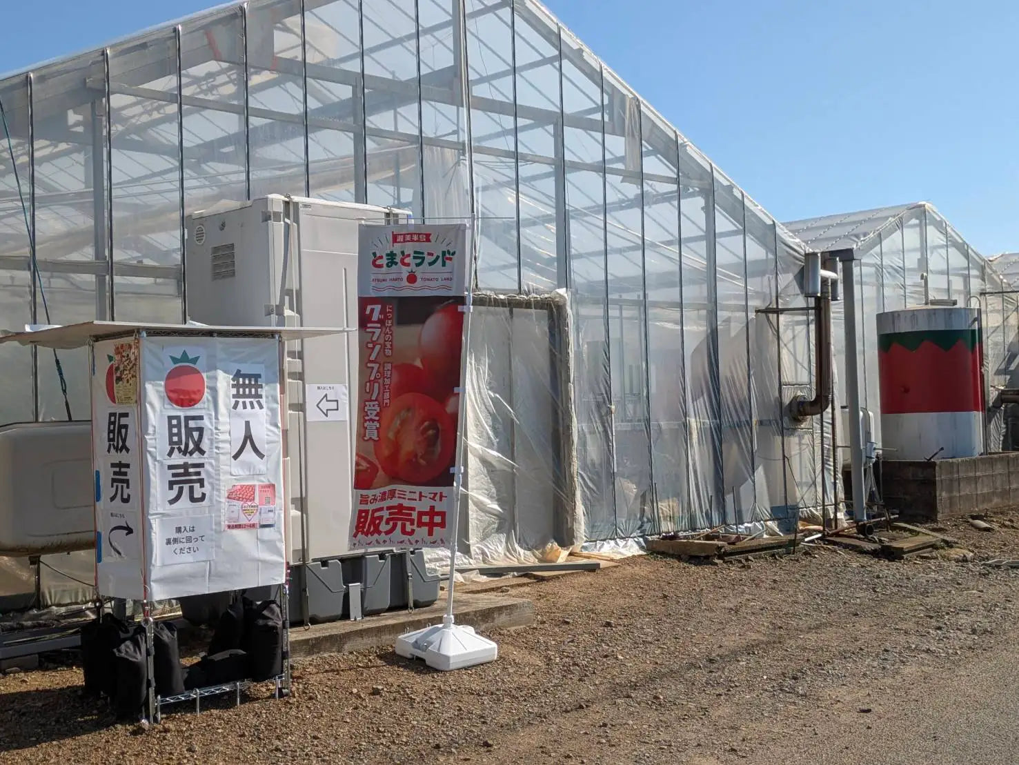 青空の下、ビニールハウスの外に設置された「とまとランド」の無人販売所。グランプリ受賞を伝えるのぼりと「無人販売」の案内板が立つ