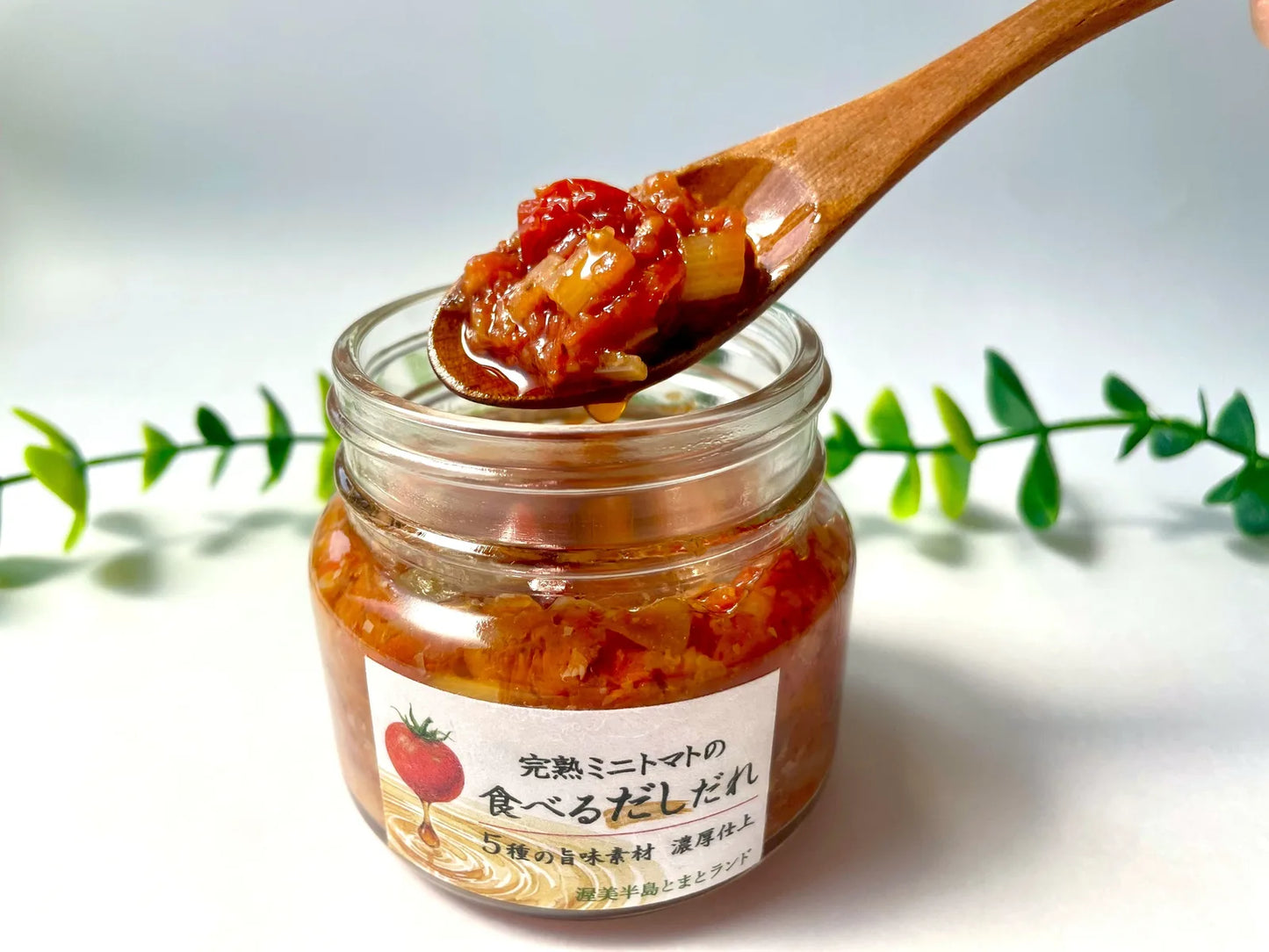 「完熟ミニトマトの食べるだしだれ」の瓶から木のスプーンで具だくさんのトマトソースをすくい上げている写真。背景に観葉植物