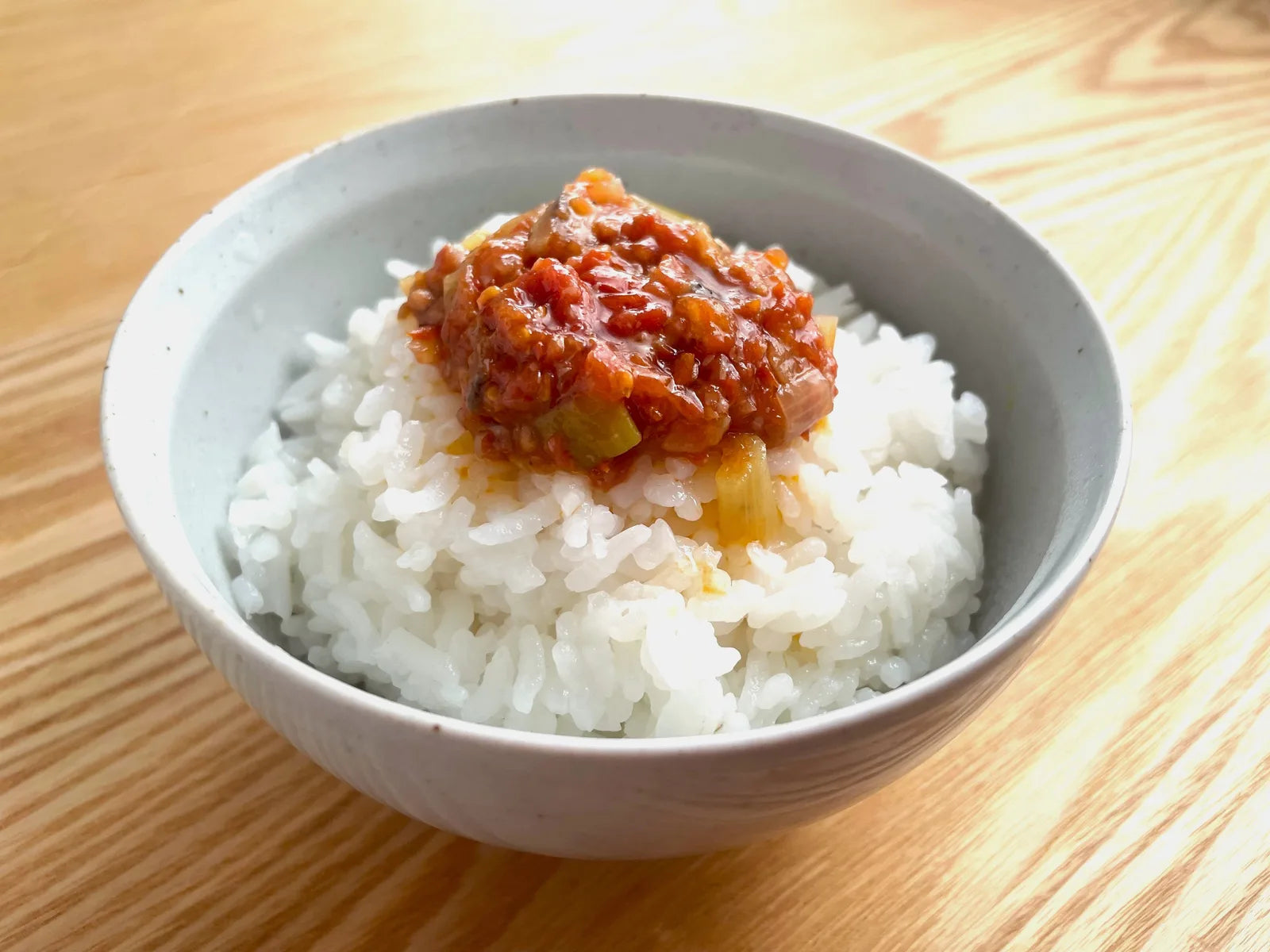 白いご飯の上に完熟ミニトマトの食べるだしだれをのせた茶碗の写真。木のテーブルの上で撮影された、ご飯のお供としての食べ方提案