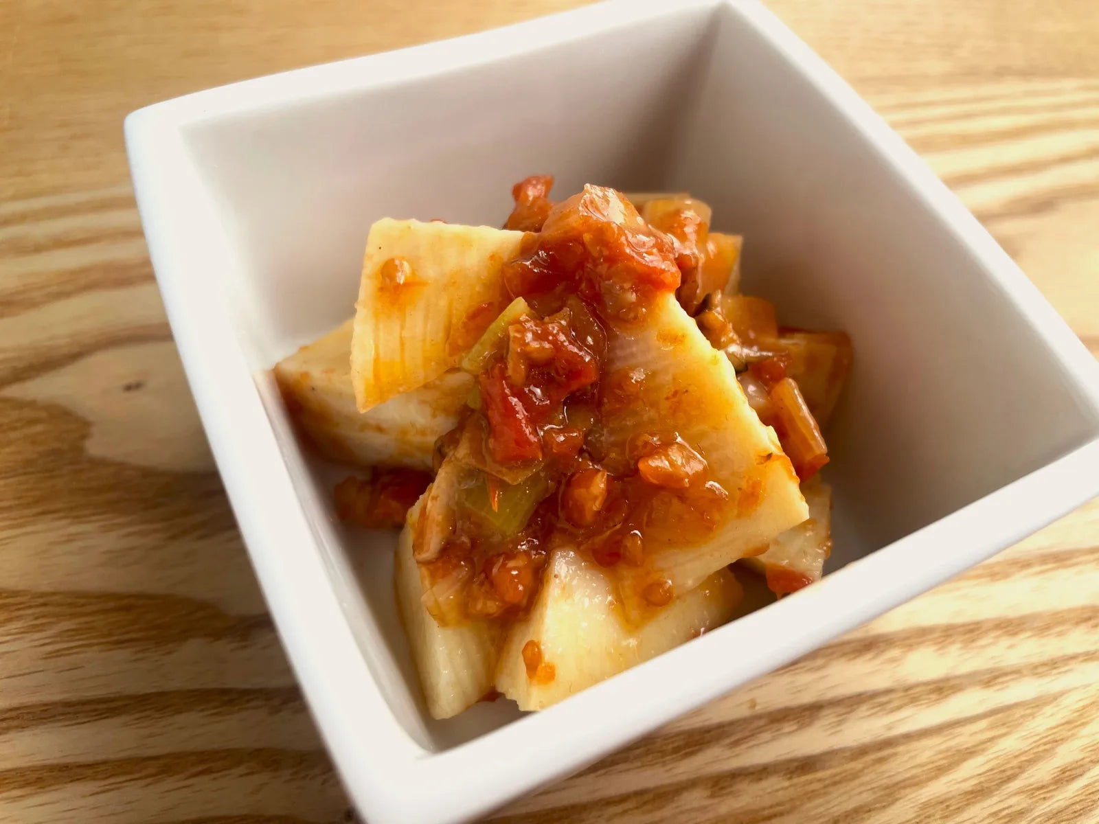 白い四角い器に盛られたたけのこの煮物に、完熟ミニトマトの食べるだしだれをのせた料理写真。木のテーブルで撮影