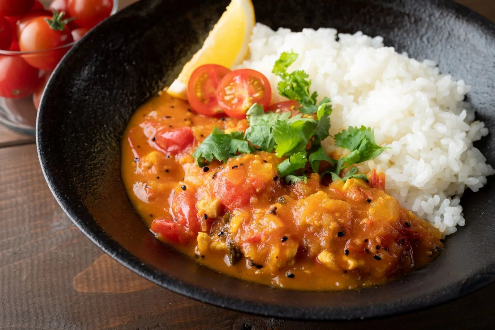 黒い器に盛られたカレーライスの写真。ミニトマトのスライスとパクチー、レモンが添えられ、背景にミニトマトの器が写る