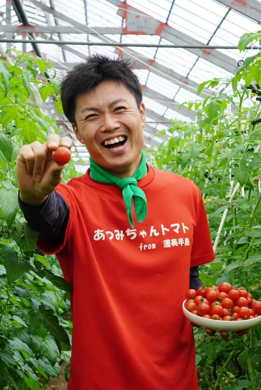 赤いTシャツに緑のスカーフの男性がハウス内でミニトマトを一粒手に持ち、もう片方の皿には収穫したミニトマトを山盛りにして笑顔で見せている写真
